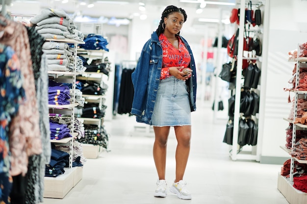 Moderna mujer afroamericana con camisa roja y falda de jeans con chaqueta posada en la tienda de ropa Es hora de ir de compras