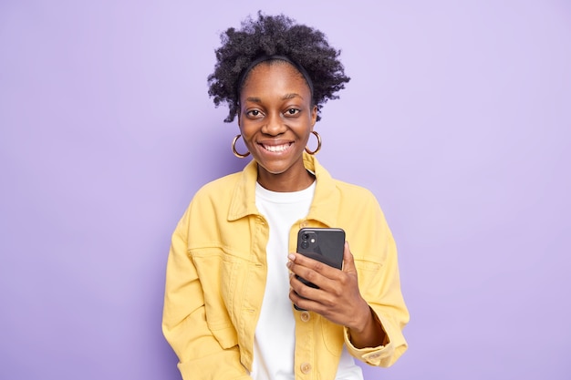 Moderna mujer afroamericana de piel oscura con cabello rizado natural chats en el teléfono utiliza el teléfono móvil tiene smartphone sonríe agradablemente