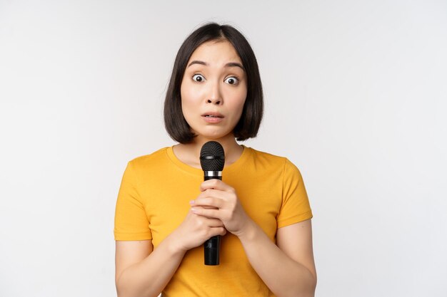 Modesta chica asiática sosteniendo el micrófono asustado hablando en público de pie contra el fondo blanco Copiar espacio