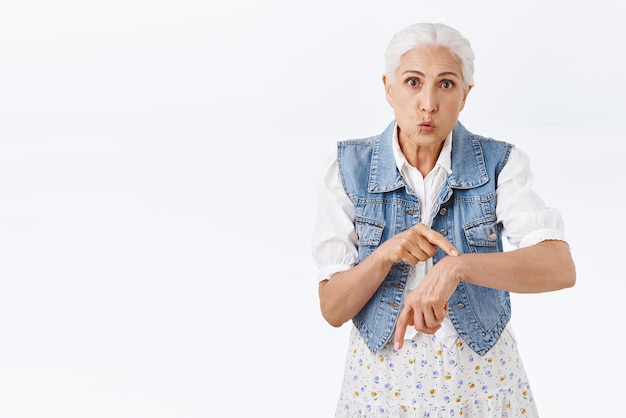 Molesta anciana enojada indignada porque el marido llega tarde señalando la muñeca como si indicara un reloj mirando la cámara furiosa quejándose de que alguien se tomó demasiado tiempo parado en el fondo blanco