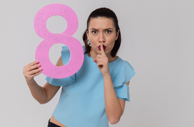 Molesta bastante joven sosteniendo rosa número ocho y haciendo gesto de silencio