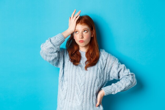 Molesta y cansada adolescente pelirroja pone los ojos en blanco, con la palma de la mano y suspirando molesto, de pie en suéter contra el fondo azul.