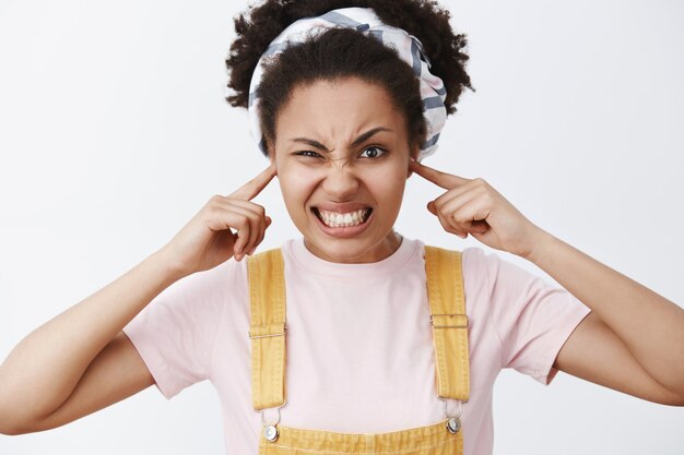 Molesta y disgustada linda estudiante afroamericana con estilo en monos amarillos y diadema, cerrando las orejas con los dedos, frunciendo el ceño, apretando los dientes