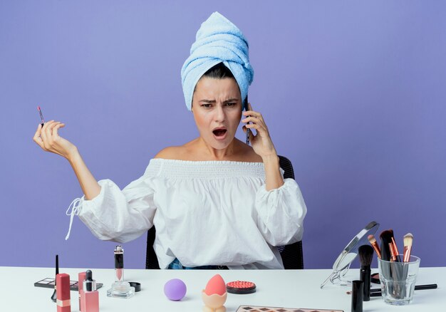 Molesta hermosa chica envuelta en una toalla para el cabello se sienta en la mesa con herramientas de maquillaje con brillo de labios hablando por teléfono aislado en la pared púrpura