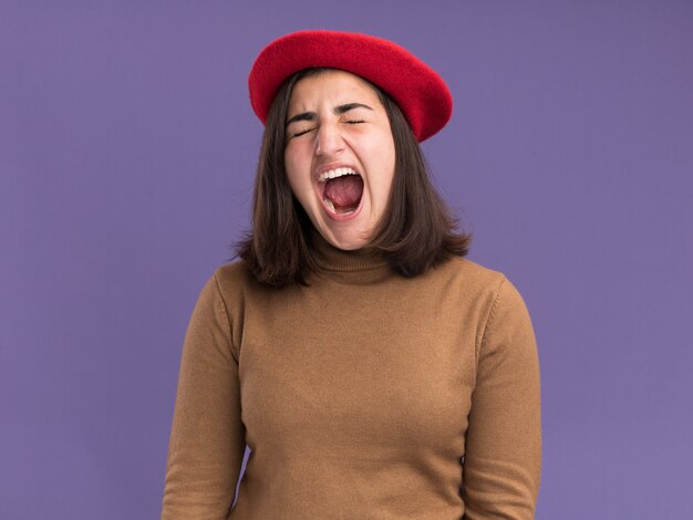 Molesta joven bastante caucásica con sombrero de boina se encuentra con los ojos cerrados gritando a alguien aislado en la pared púrpura con espacio de copia