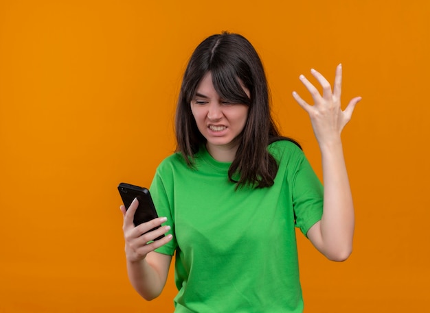 Molesta joven caucásica en camisa verde sostiene el teléfono y levanta la mano sobre fondo naranja aislado