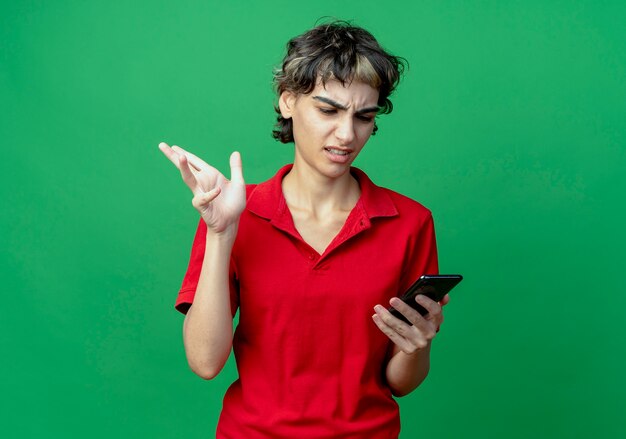 Molesta joven caucásica con corte de pelo pixie sosteniendo y mirando el teléfono móvil manteniendo la mano en el aire aislado sobre fondo verde con espacio de copia