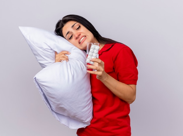 Molesta joven caucásica enferma abrazando la almohada poniendo la cabeza sobre ella con un paquete de tabletas y un vaso de agua en la mano mirándolos aislado sobre fondo blanco con espacio de copia