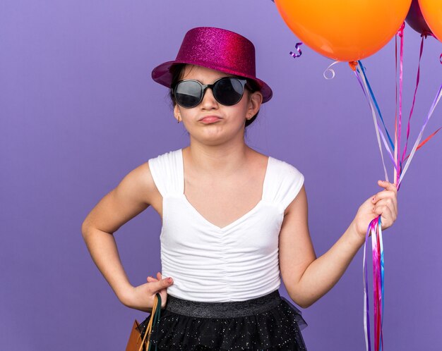 Molesta joven caucásica en gafas de sol con sombrero de fiesta violeta sosteniendo globos de helio y bolsas de compras aisladas en la pared púrpura con espacio de copia