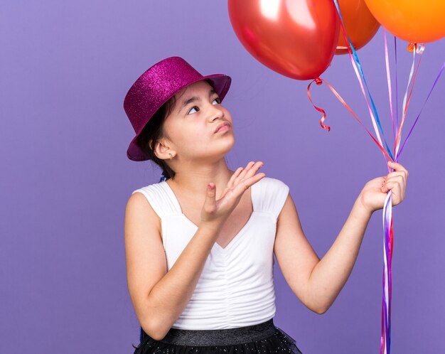 Molesta joven caucásica con gorro de fiesta violeta sosteniendo y apuntando a globos de helio aislados en la pared púrpura con espacio de copia