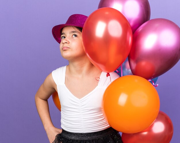 Molesta joven caucásica con gorro de fiesta violeta sosteniendo globos de helio y mirando hacia arriba aislado en la pared púrpura con espacio de copia