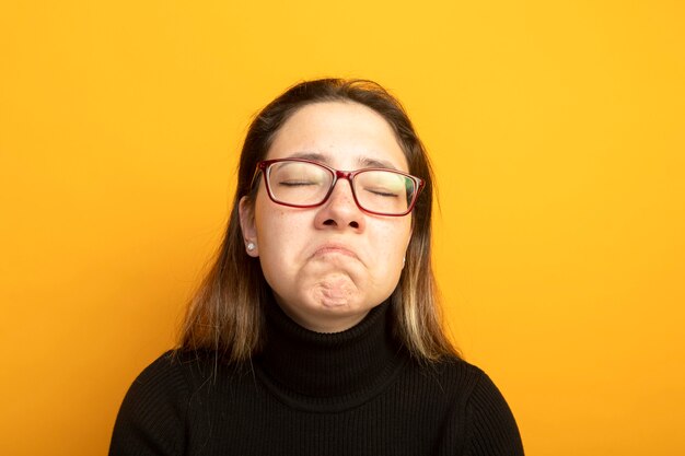 Molesta joven hermosa en un cuello alto negro y gafas con los ojos cerrados