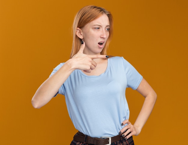 Molesta joven pelirroja pelirroja con pecas mirando y apuntando al lado en naranja