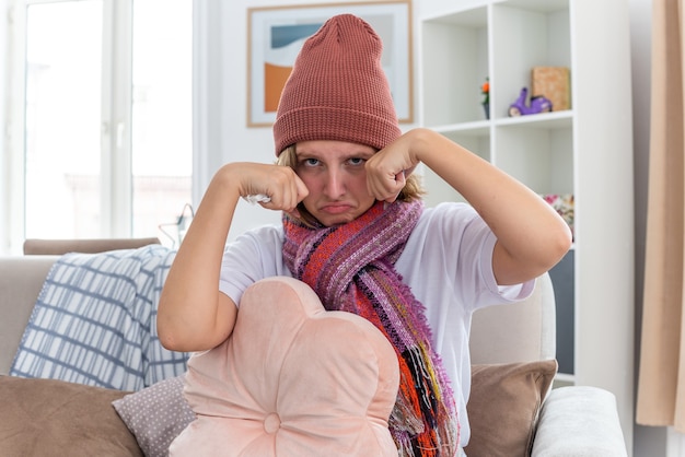 Foto gratuita molesta mujer joven insalubre con gorro con bufanda con aspecto de malestar y enfermo que sufre de resfriado y gripe llorando duro frotándose los ojos sentado en el sofá en la sala de estar luminosa