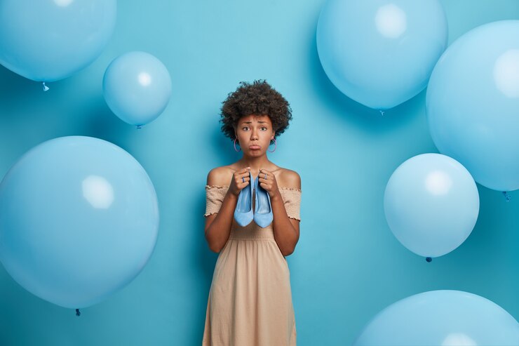 Foto gratuito molesta mujer de piel oscura con cabello rizado sostiene zapatos de tacón azul se prepara para poses de fiesta contra globos azules.