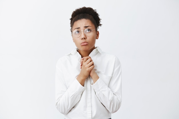 Molesta, tímida e insegura, atractiva chica afroamericana con gafas y camisa de cuello blanco, apretando las palmas de las manos y frunciendo el ceño, mirando con tristeza y expresión miserable sobre la pared gris