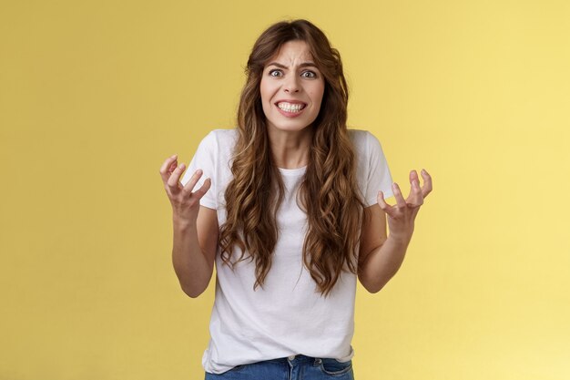 Molesto caucásico de pelo rizado irritado niña angustiada encogerse indignado apretar los dientes fruncir el ceño furioso puños de sqeez quejándose de odio argumentando que luchan molestos presionado tienen disputa fondo amarillo.