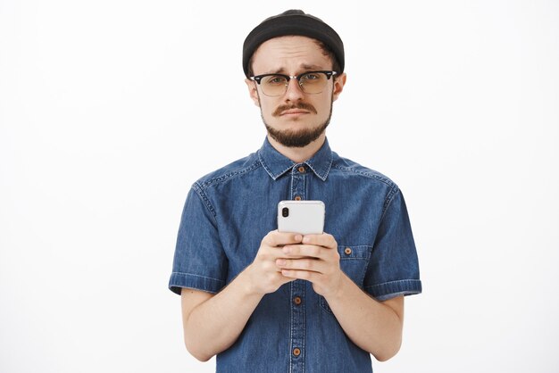 Molesto chico barbudo lindo con gorro negro y gafas haciendo cara triste y frunciendo el ceño sosteniendo un teléfono inteligente expresando celos o arrepentimiento por perder la oportunidad de comprar boletos en línea