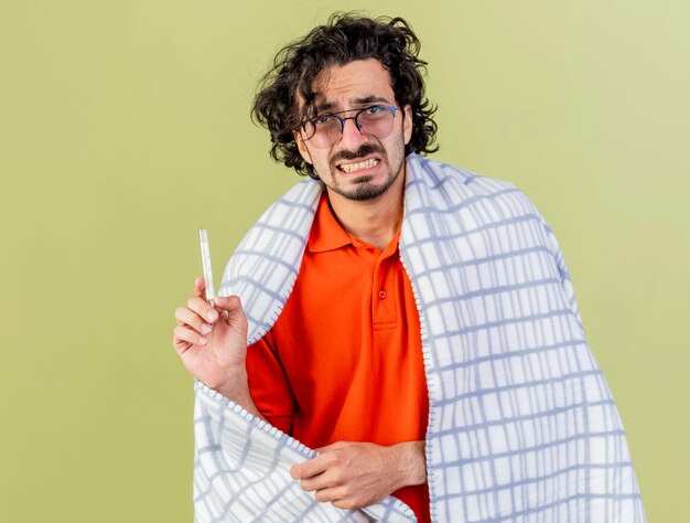 Molesto joven enfermo con gafas envuelto en plaid sosteniendo un termómetro agarrando plaid mirando al frente aislado en la pared verde oliva