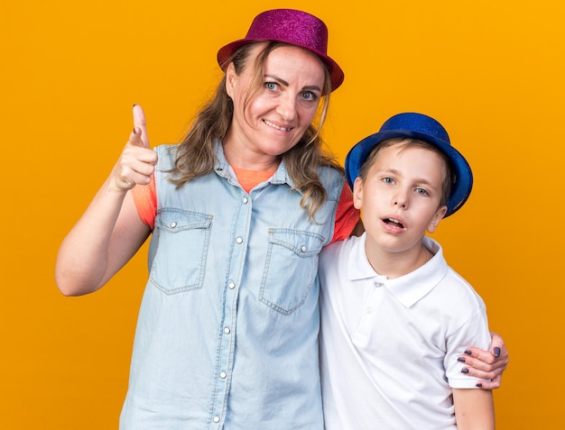 Foto gratuita molesto joven eslavo con gorro de fiesta azul de pie con su madre vistiendo gorro de fiesta púrpura y apuntando al lado aislado en la pared naranja con espacio de copia