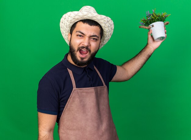 Molesto joven jardinero varón caucásico vistiendo sombrero de jardinería sosteniendo maceta aislado en la pared verde con espacio de copia