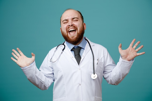 Molesto joven médico masculino con abrigo médico y estetoscopio alrededor del cuello mostrando las manos vacías gritando con los ojos cerrados aislados en el fondo azul