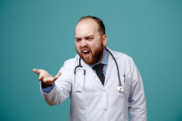 Molesto joven médico masculino con abrigo médico y estetoscopio alrededor de su cuello mirando al costado mostrando la mano vacía aislada en el fondo azul