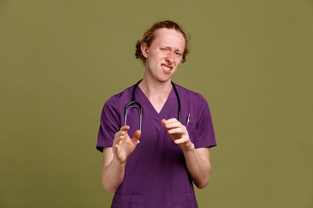molesto joven médico masculino vistiendo uniforme con estetoscopio aislado sobre fondo verde