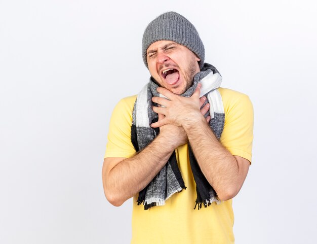 Molesto joven rubio enfermo eslavo vestido con gorro de invierno y bufanda se ahoga con las manos aisladas en la pared blanca con espacio de copia