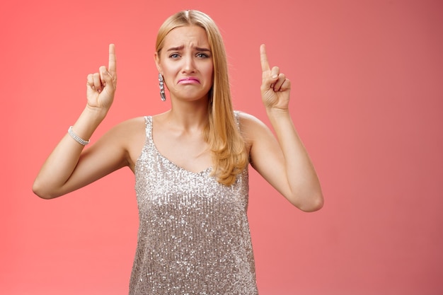 Foto gratuita molesto lloriquear inmadura mimada rubia rica chica en vestido plateado brillante haciendo pucheros frunciendo el ceño va a llorar apuntando hacia arriba arrepentimiento celos pidiendo comprar zapatos caros, de pie fondo rojo disgustado.