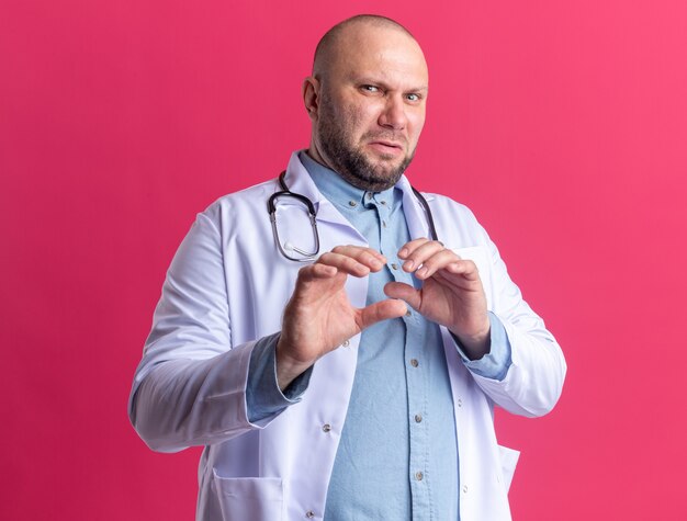 Molesto médico varón de mediana edad vistiendo una bata médica y un estetoscopio mirando al frente haciendo gesto de rechazo aislado en la pared rosa