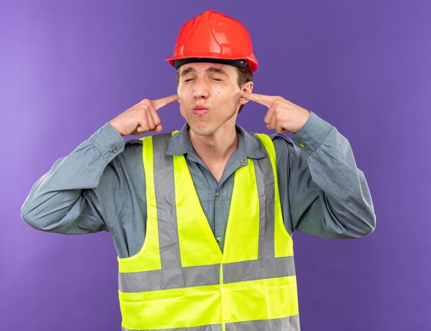 Molesto con los ojos cerrados el joven constructor en uniforme cerró los oídos aislados en la pared azul