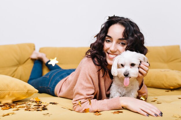 Momentos domésticos divertidos de mujer joven feliz escalofriante en el sofá en casa con mascota doméstica. Divirtiéndose, oropeles dorados, sonriente, estado de ánimo alegre, increíbles emociones positivas verdaderas