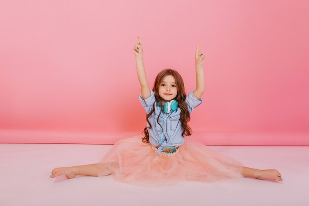 Momentos dulces infancia feliz de increíble joven en falda de tul haciendo qymnastics split en piso blanco sobre fondo rosa. Lindo niño de moda con cabello largo morena, cabezas azules en el cuello