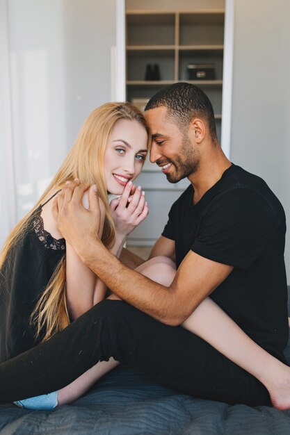 Momentos de la encantadora pareja de chico guapo cuidando a una joven bonita con cabello largo y rubio, abrazándola en la cama en un apartamento moderno. Relajándonos juntos por la mañana, amor, relación