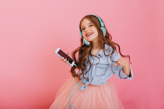 Momentos encantadores de niño disfrutando de la música a través de auriculares, bailando con teléfono aislado sobre fondo rosa. Expresando verdaderas emociones positivas del niño feliz de moda en el entretenimiento