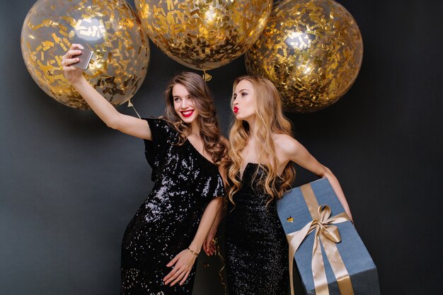 Momentos de fiesta feliz de dos mujeres jóvenes de moda haciendo selfie. Vestido negro de lujo, cabello largo y rizado, globos grandes con oropeles dorados, presente, divirtiéndose, sonriendo.