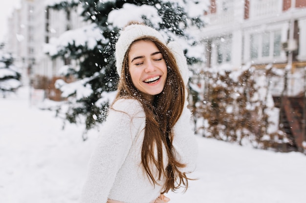 Momentos de invierno feliz de mujer joven alegre con cabello largo morena, ropa de invierno blanca divirtiéndose en la calle en tiempo de nevando. Expresando positividad, verdaderas emociones brillantes, sonriendo con los ojos cerrados.