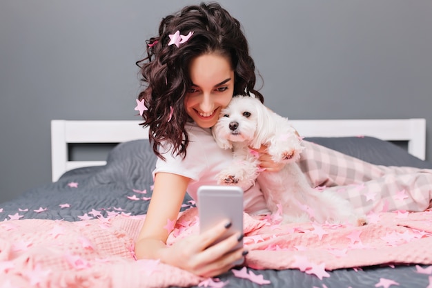 Momentos lindos felices en casa con mascotas de hermosa mujer morena en pijama. Haciendo selfie en la cama en oropel rosa, sonriendo, expresando felicidad