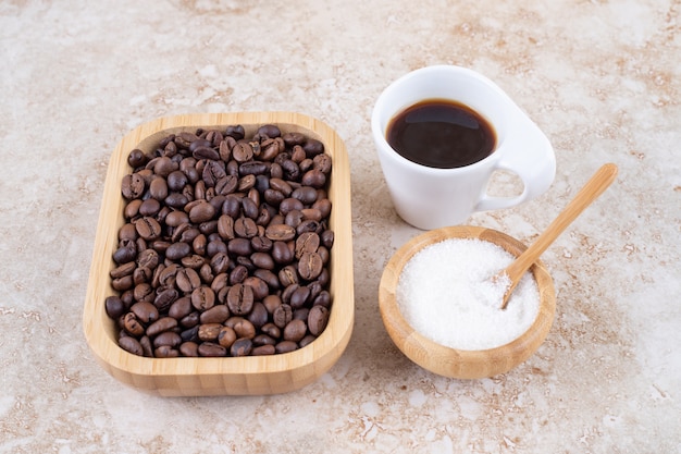 Montón de granos de café en una bandeja de madera junto a un tazón pequeño de azúcar y una taza de café