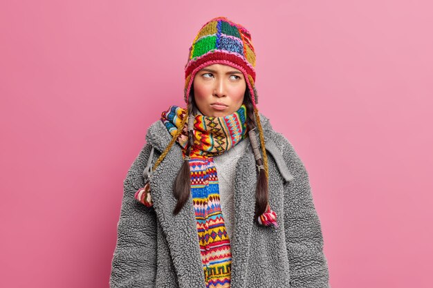 Moody mujer asiática disgustada concentrada en algún lugar con expresión ofendida lleva bufanda de punto y abrigo de piel gris posa contra la pared rosa del estudio