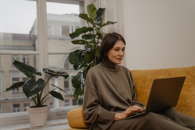 Morena adulta caucásica sosteniendo una computadora portátil moderna de rodillas mientras se sienta en un sofá en la habitación durante el día Concepto de navegación en la tienda en línea