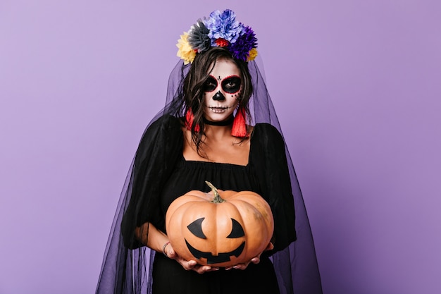 Morena con arte facial de calavera mexicana sostiene calabaza decorada. Foto interior de niña con corona de flores.