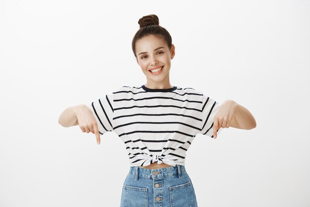 Foto gratuita morena atractiva alegre sonriendo ampliamente, señalando con el dedo hacia abajo