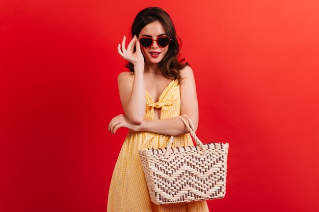 Morena elegante en vestido amarillo se pone gafas en forma de corazones. Chica con bolsa de playa posando en la pared roja.