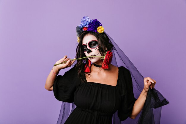 Morena con flores en el pelo se ve con picardía, posando con rosa roja. Retrato de mujer con máscara de calavera y velo negro.