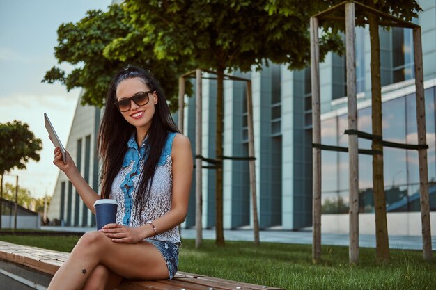 Una morena hermosa y feliz con un tatuaje en la pierna, con ropa de moda y gafas de sol, sostiene un café para llevar y una tableta digital mientras se sienta en un banco contra un rascacielos, mirando un came