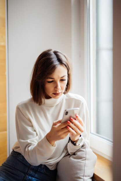 Morena interesada bonita mujer adolescente en camiseta blanca con mensajes de texto de teléfono inteligente