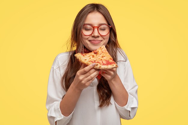 Morena joven hambrienta con gafas posando contra la pared amarilla