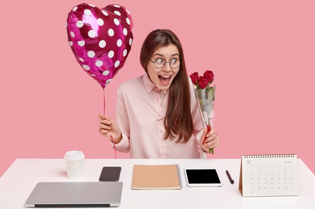 Morena joven mujer caucásica sosteniendo globos en forma de corazón y ramo de rosas rojas